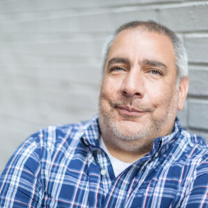 Paul Marengo has light skin and a closely shaven beard and hair with grey and black hair. Paul wears a blue plaid shirt and in photographed in front of a white brick wall.