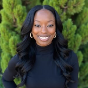 AliShah Watson has medium brown skin and long wavy hair. AliShah wears a long sleeve black turtleneck and gold and pearl earrings and is photographed in front of green foilage.