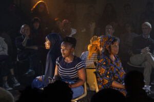 Four women sit on chairs in a circle backs to each other. Some wear traditional South African clothing and some are dressed in modern attire. Around them an audience can be seen watching.