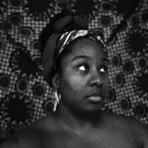 This is a black and white image. Lisa Brown has brown skin and hair that is pulled up in a wrapped scarf. Lisa's shoulders are bare and they are photographed in front of a patterned background.