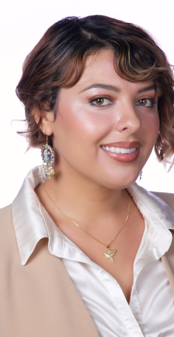 Celestina Billington has medium light skin and short wavy brown hair. Celestina is wearing a white collared blouse, tan jacket, and dangling earrings.