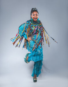 Nomfusi is shot in a studio against a white background. The artist wears a turquoise jumpsuit adorned with colorful fabric strips.