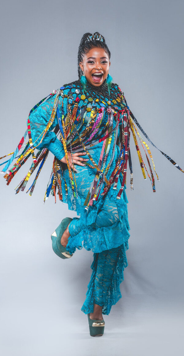 Nomfusi is shot in a studio against a white background. The artist wears a turquoise jumpsuit adorned with colorful fabric strips.