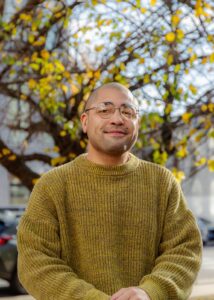 Zack Blackwood has light olive skin and a closely shaven head. Zach wears a heathered lime green sweater and round glasses. They are photographed in front of a tree.