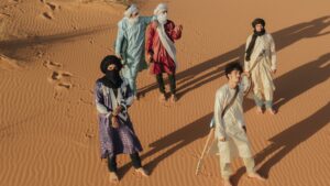 The five members of Tarwa N-Tiniri are dressed in traditional Moroccan clothing and photographed walking across dessert sands. Some hold their instruments.