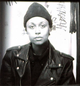 This is a black and white image. Shawné Michaelain Holloway is photographed in front of a wall with graffiti-like writing. They wear a dark shirt, dark leather jacket, and dark beanie cap.