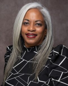 Jamila Jones has medium brown skin and long, grey, straight hair. Jamila wears a black and white patterned top and is photographed against a grey background.