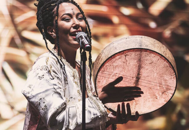 Artist The Leila plays a flat frame drum while singing at a stand microphone. They are on a brightly lit stage. The Leila wears an intricately embroidered white jacket and their hair in long braids.