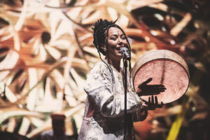 Artist The Leila plays a flat frame drum while singing at a stand microphone. They are on a brightly lit stage. The Leila wears an intricately embroidered white jacket and their hair in long braids.