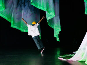 A dancer with arms raised jumps diagonally across a stage. Green and blue curtains blow behind.